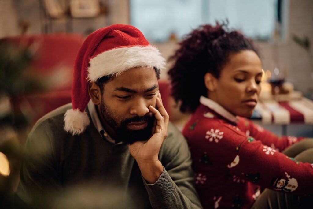 A Person Experiencing Grief At Christmas, Sitting Quietly In A Holiday Setting With A Partner Nearby, Reflecting The Emotional Weight The Season Can Bring.